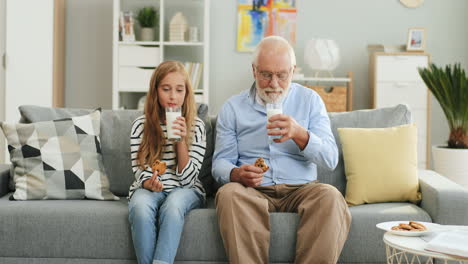 Hübsches-Blondes-Jugendlich-Mädchen,-Das-Zusammen-Mit-Ihrem-Großvater-Mit-Grauen-Haaren-Auf-Dem-Sofa-Zu-Hause-Sitzt-Und-Milch-Mit-Keksen-Trinkt.